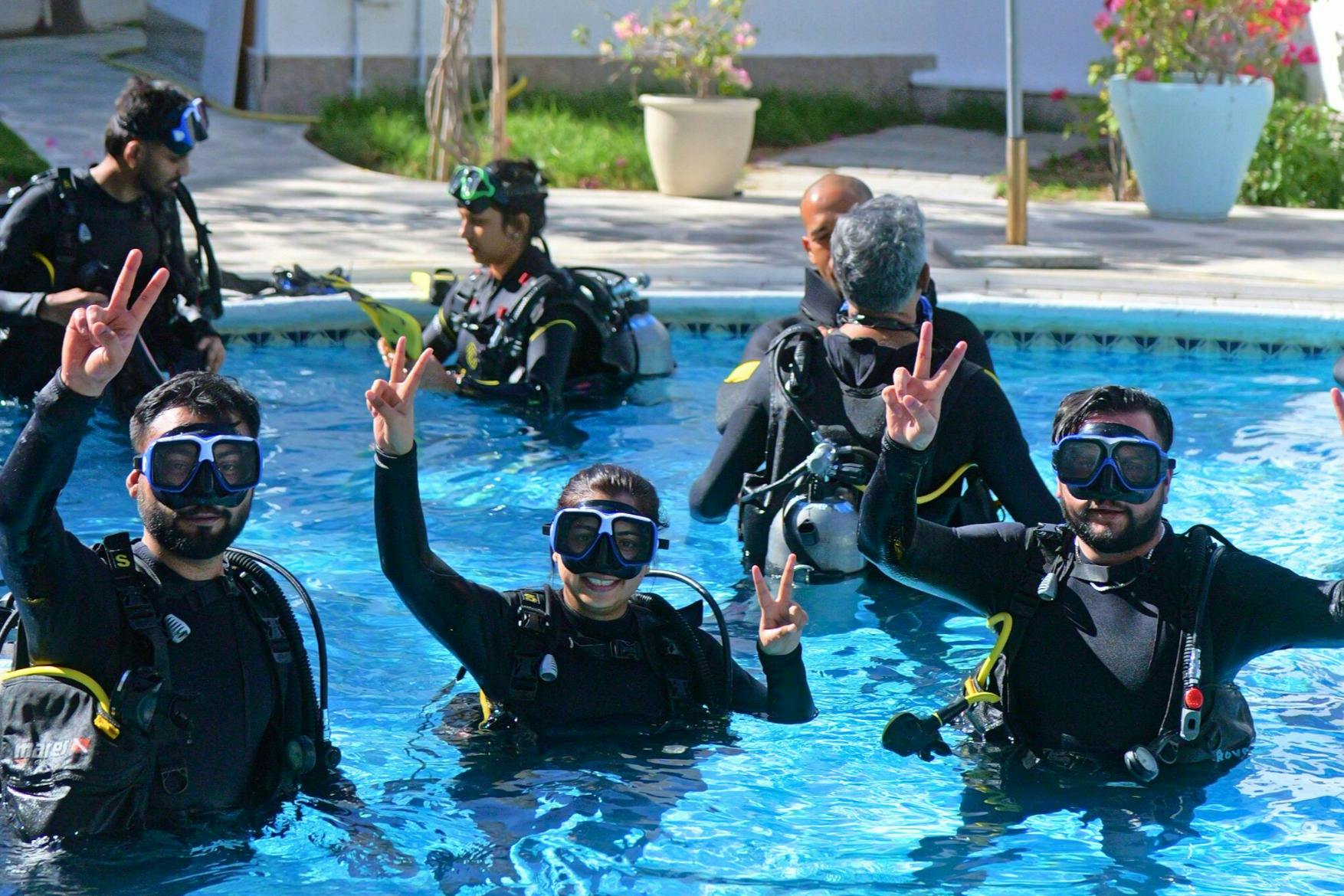 Zwei Taucher in einem Schwimmbecken mit voller Ausrüstung, die lächeln und Friedenszeichen machen, während andere im Hintergrund üben.
