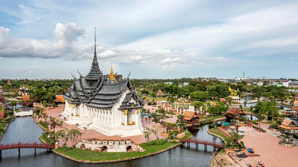 Aerial view of a traditional Thai temple with ornate roofs, surrounded by trees, bridges, and water on a sunny day.