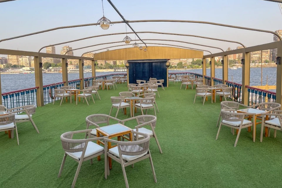 An outdoor seating area on a boat with green artificial grass flooring, tables, chairs, and a partial cityscape in the background.