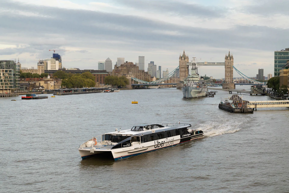 A boat labeled 'Uber Boat' on the Thames River with Tower Bridge and city buildings in the background.