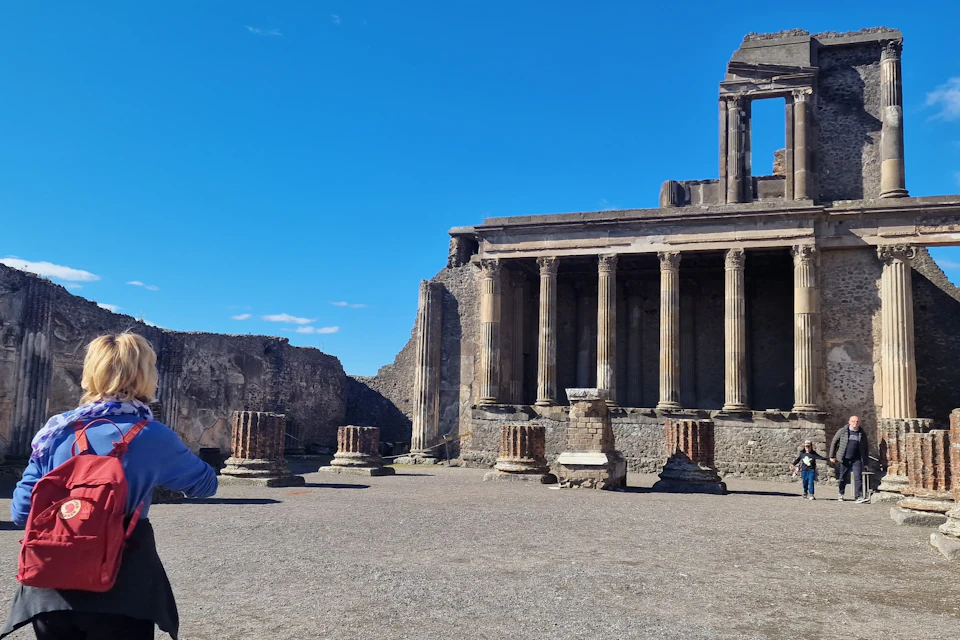 Ancient ruins featuring columns and a partially standing building under a clear blue sky, with a person on the left side of the scene.