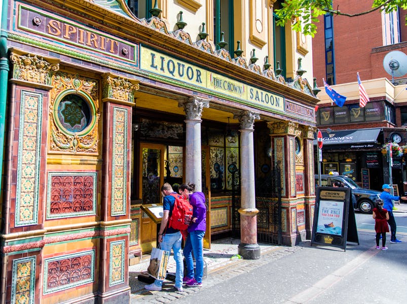 Three people stand outside a colorful, ornate pub called "The Crown Saloon," reading a street map. Adjacent shops and flags are visible.