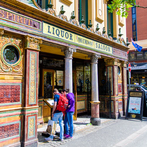 Tre personer står utanför en färgglad, utsmyckad pub som heter "The Crown Saloon" och läser en gatukarta. Angränsande butiker och flaggor är synliga.