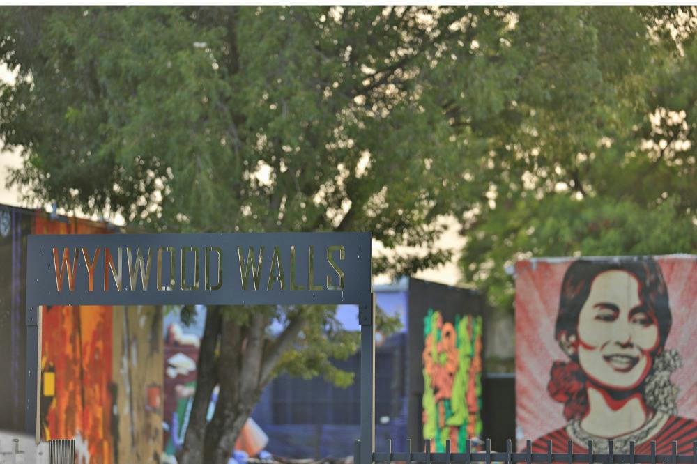Outdoor scene featuring a mural of a smiling woman with colorful artwork and trees in the background, and a sign reading 'WALLS.'