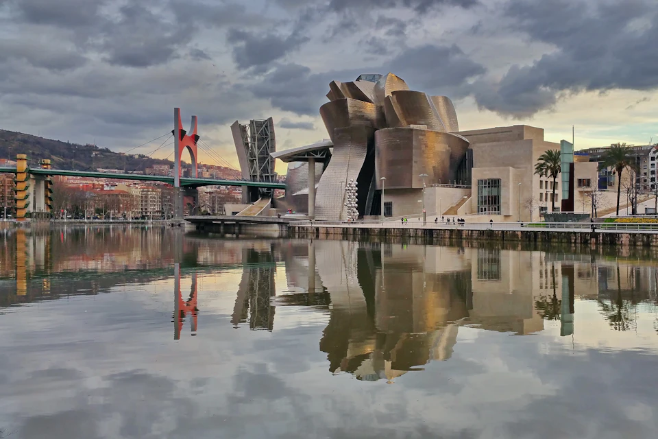 Modern building with metallic, abstract architecture reflected in a calm river, with a red and green bridge in the background.