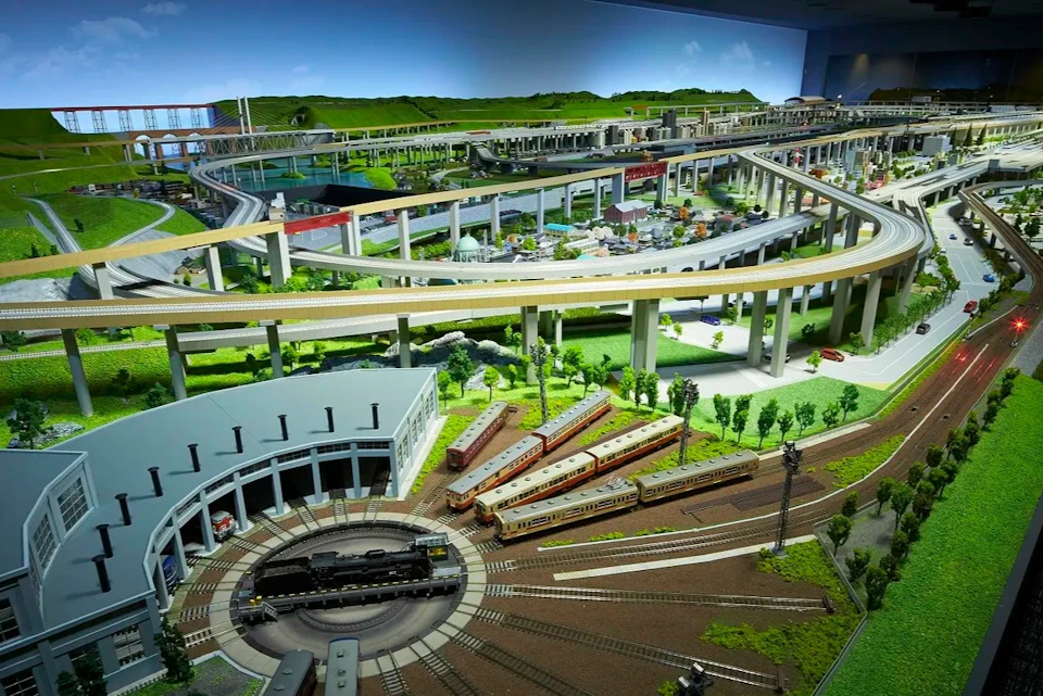 A detailed model train layout featuring a roundhouse, multiple tracks, trains, and elevated tracks over a landscaped area.