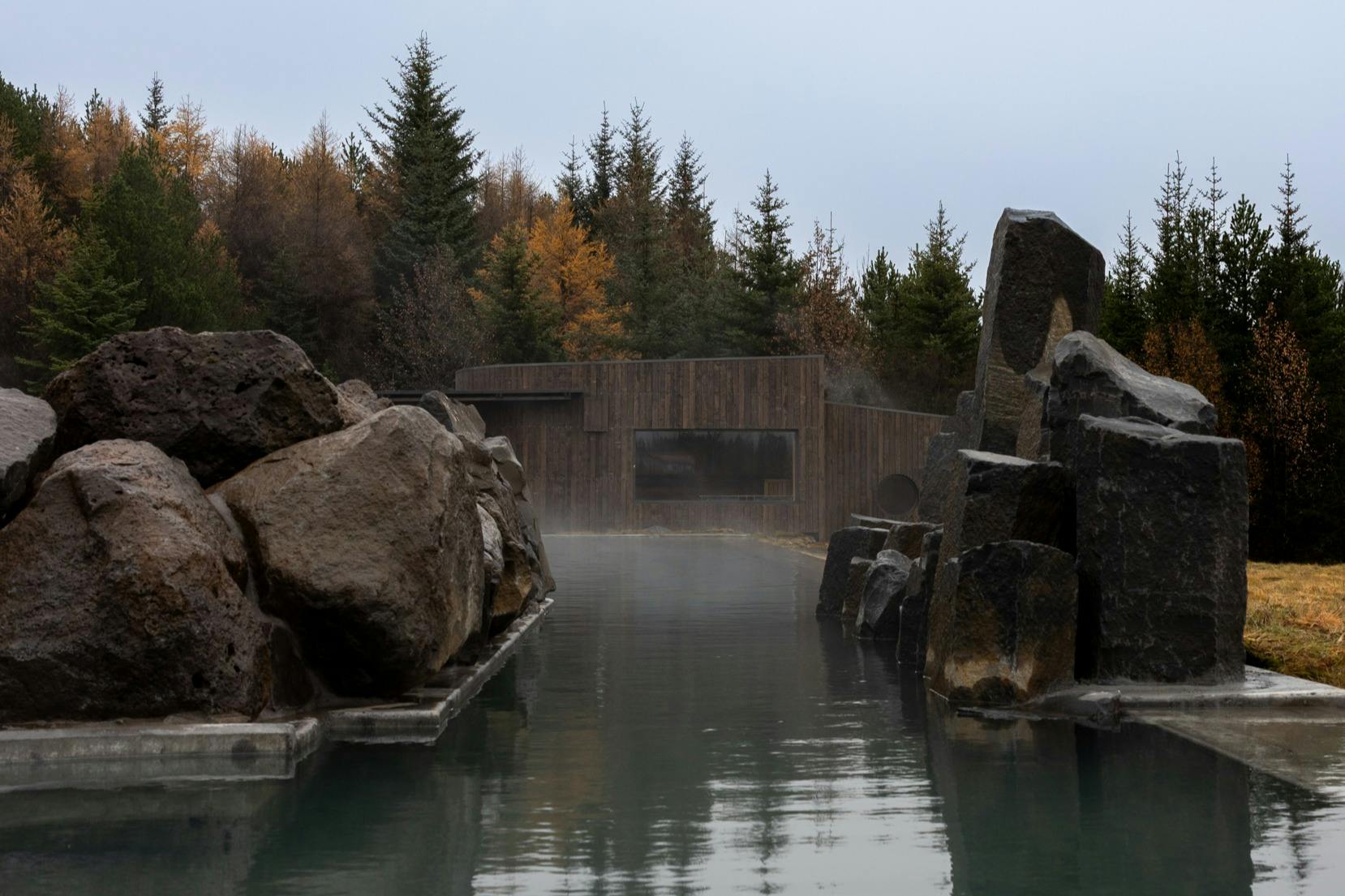 A steaming, narrow hot spring surrounded by large rocks, leading to a wooden structure with a backdrop of autumn trees and an overcast sky.