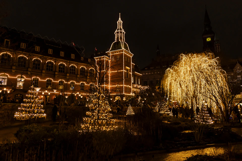 A building with a tower and trees are adorned with warm Christmas lights at night, creating a festive atmosphere.