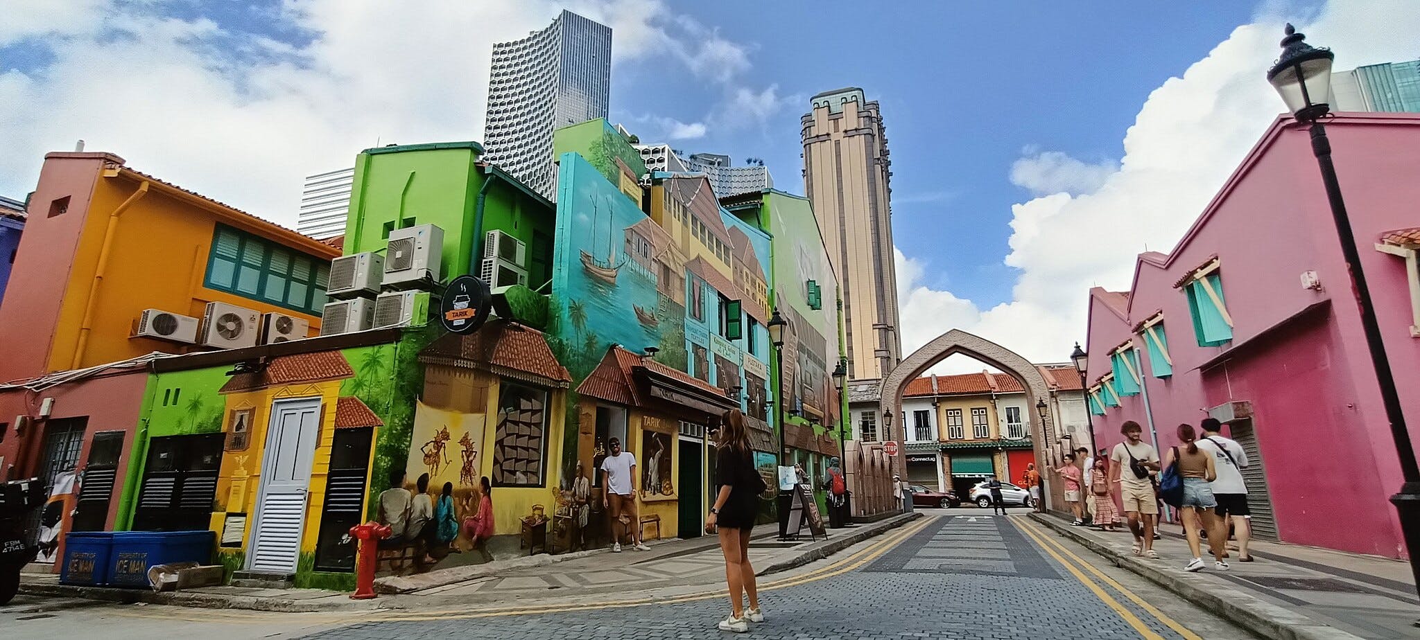 Edifici colorati con murales, condizionatori d'aria e persone che camminano su una strada di ciottoli sotto un cielo azzurro.