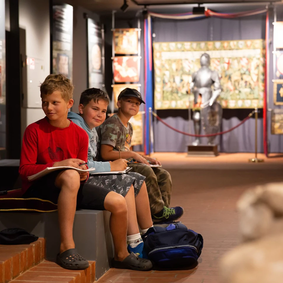 Three boys sit on a bench inside a museum, sketching. Behind them are historical exhibits, including a suit of armor.