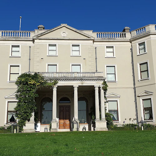 En stor, beige herrgård med många fönster, en entré, murgrönsklädda pelare och välskötta gröna gräsmattor, under en klarblå himmel.