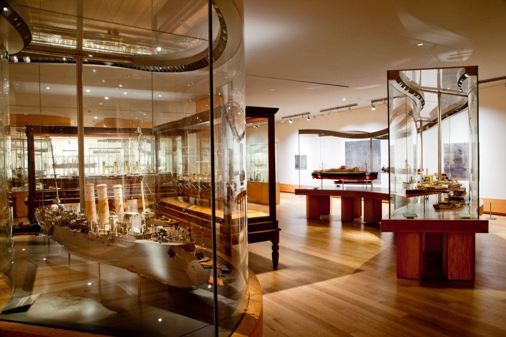 Una exposición museística con detallados modelos de barcos expuestos en vitrinas de cristal, con suelos de madera e iluminación en el techo.