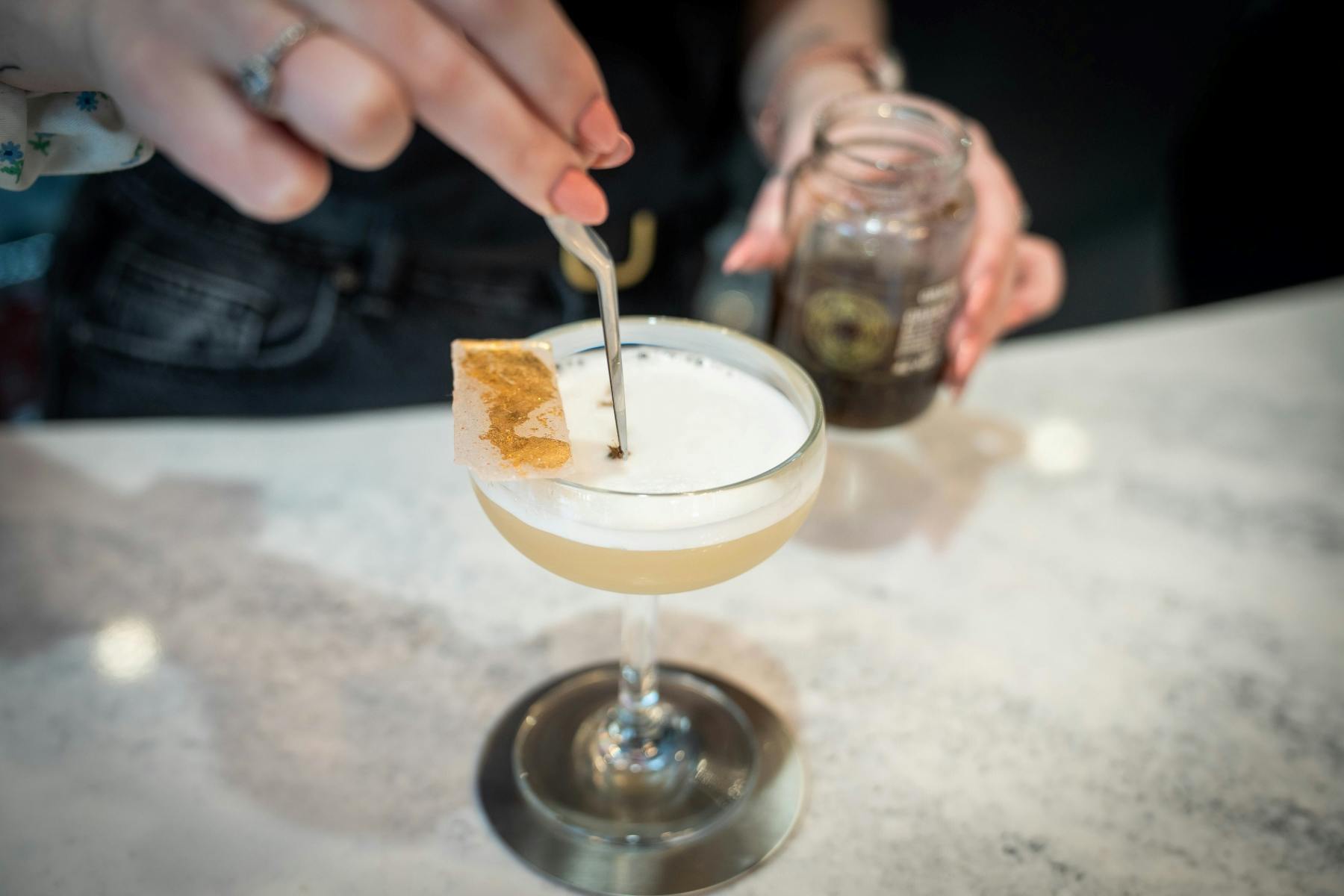 泡立ったカクテルを小さな道具でステムグラスに飾っている人が、もう一方の手に瓶を持っています。