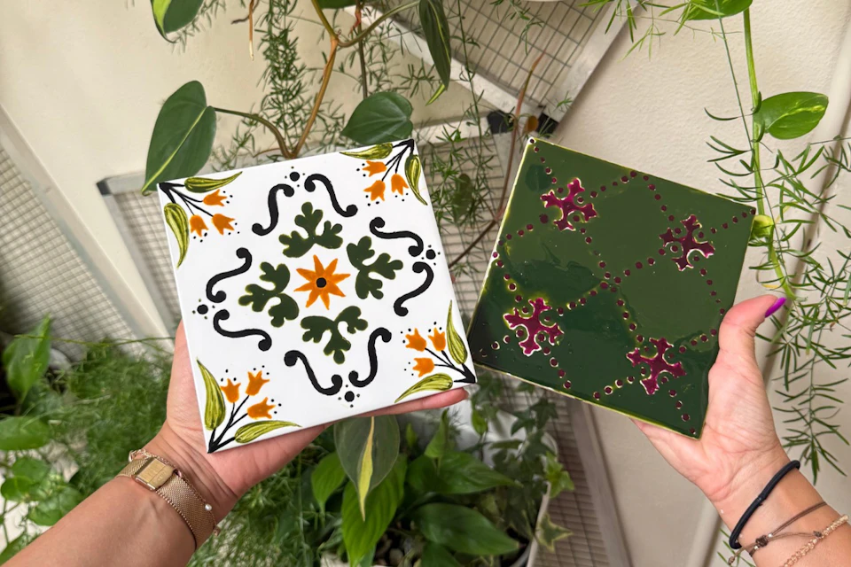 Two ceramic tiles with decorative patterns, one white with floral motifs and one green with geometric designs, held by hands.