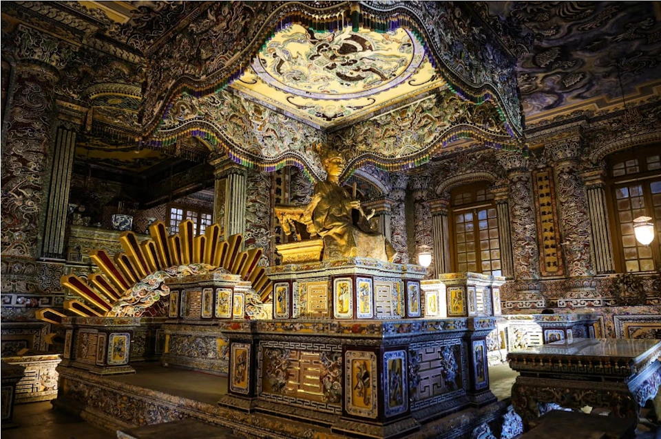 Ornate interior with a gold statue seated on an elaborately decorated pedestal, surrounded by intricate carvings and patterns.