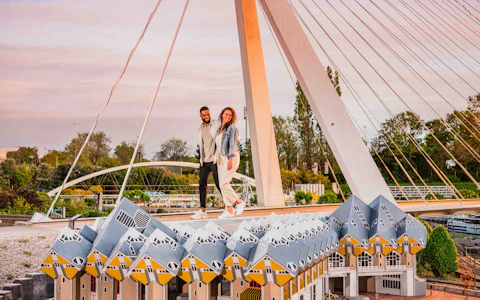 Dos personas de pie en una plataforma sobre una serie de edificios cúbicos de tejados amarillos, con un puente colgante al fondo.