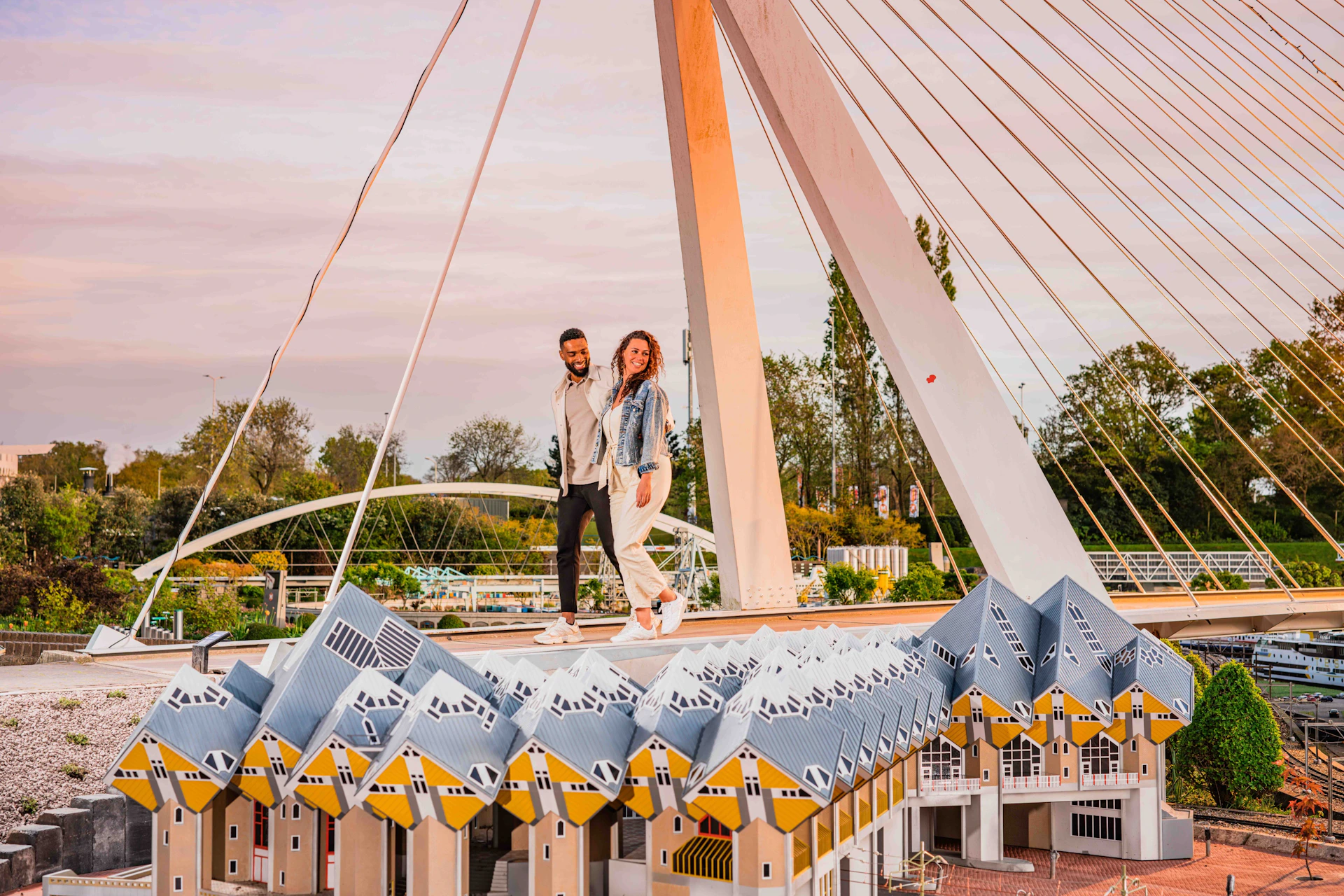 Two people standing on a platform above a series of cubic yellow-roofed buildings, with a suspension bridge in the background.