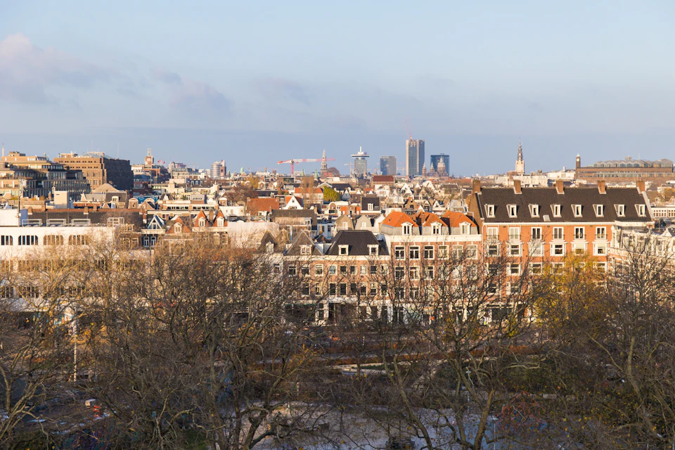 A cityscape view of Amsterdam featuring buildings, trees and cranes, set against a clear blue sky.