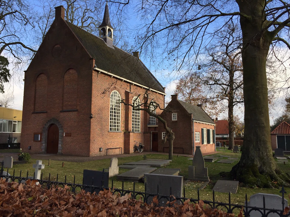 Old brick church with stained glass windows, adjacent graveyard, leafless trees, and blue sky. Some gravestones and a bike present.