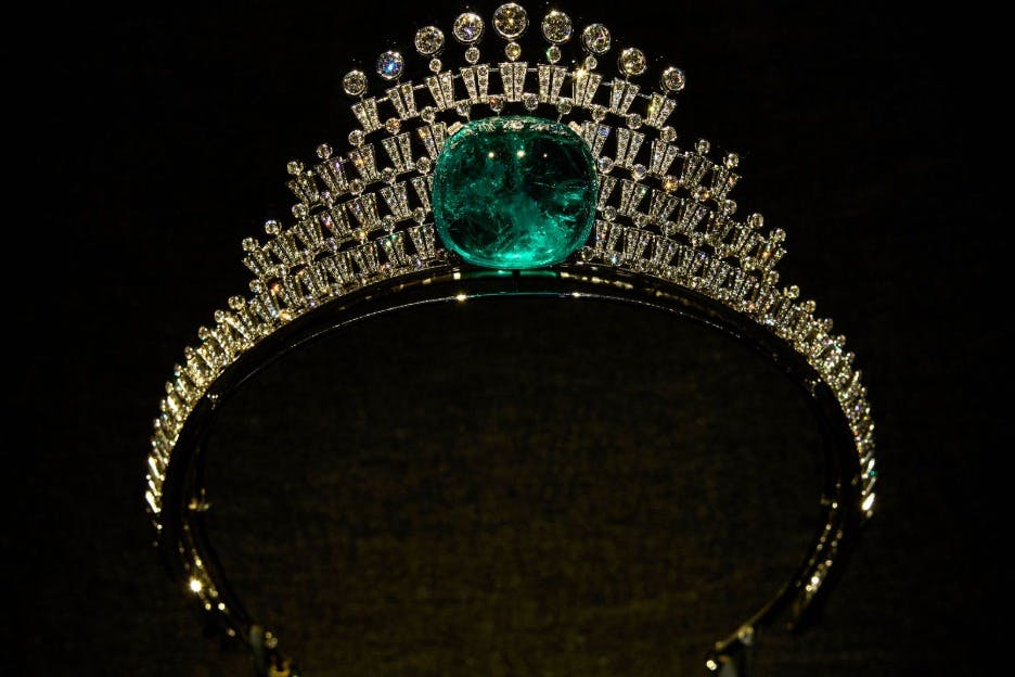 Una tiara de oro adornada con diamantes y una gran piedra preciosa verde en el centro, expuesta sobre un fondo oscuro.