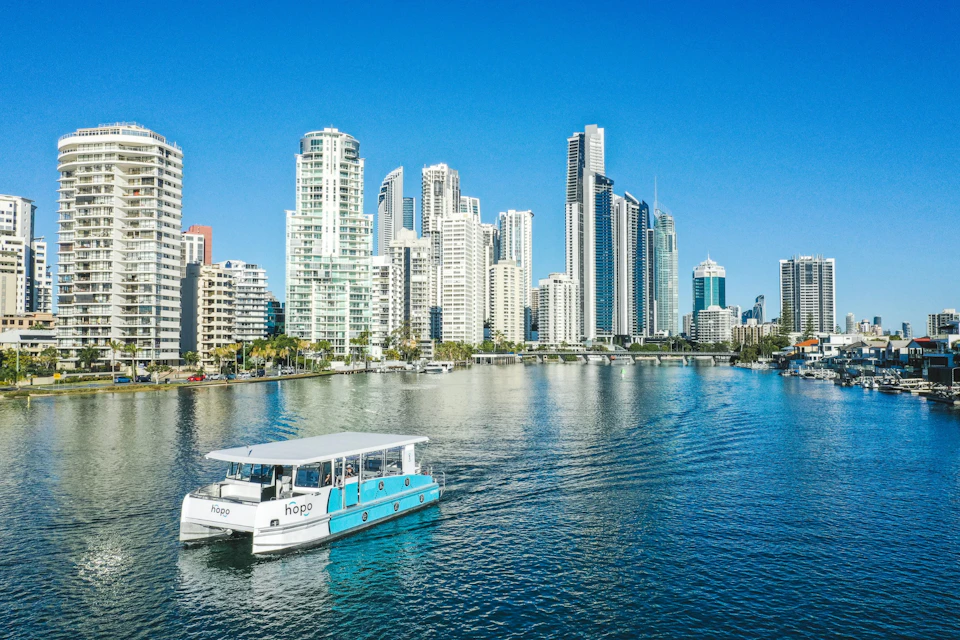 A blue and white boat on a calm river with modern city skyscrapers in the background under a clear blue sky.