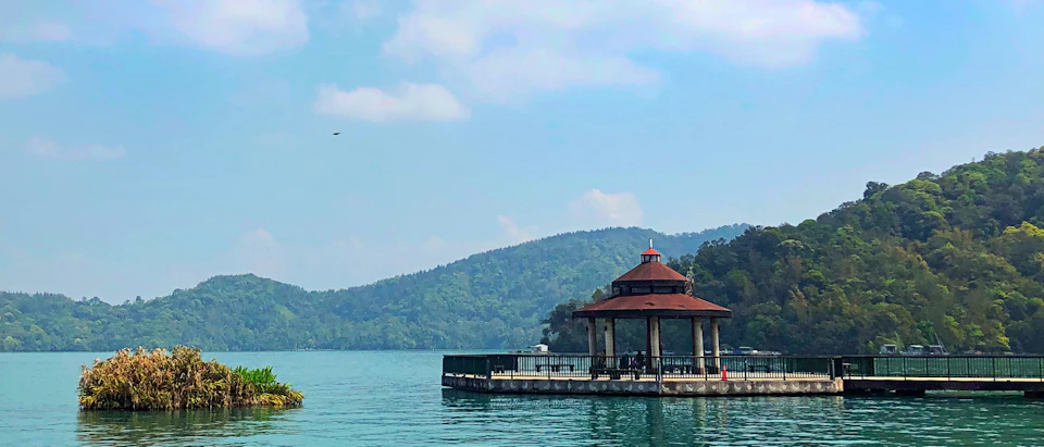A lakeside pavilion with a red roof and pillars, surrounded by calm water and backed by tree-covered hills under a clear sky.
