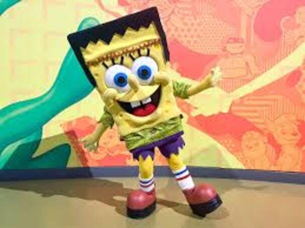 커다란 파란색 눈, 빨간색 신발, 초록색 셔츠를 입은 노란색 만화 캐릭터로 분장한 사람이 화려한 벽 앞에서 포즈를 취하고 있습니다.