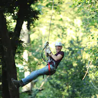 Pessoa a fazer tirolesa através de uma floresta verdejante, sorridente e com equipamento de segurança, incluindo capacete e arnês.