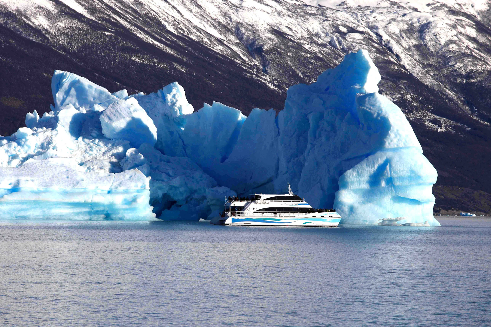 A boat is near large blue icebergs on a calm body of water, with a forested and snowy mountain in the background.