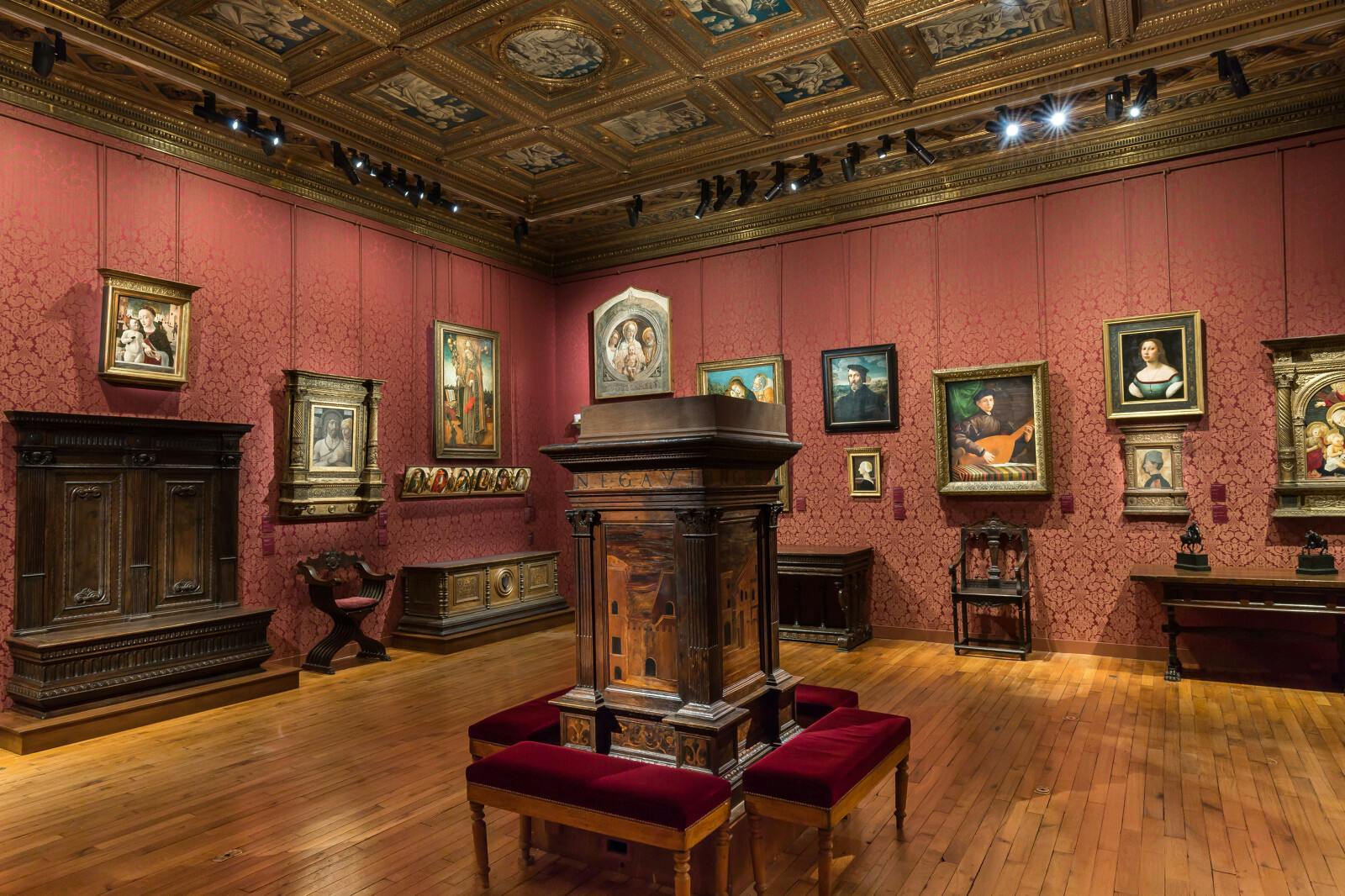 Μια γκαλερί τέχνης με κόκκινους τοίχους, ξύλινο πάτωμα και περίτεχνη οροφή. Προβάλλονται διάφορα έργα ζωγραφικής με κορνίζες και κλασικά έπιπλα.