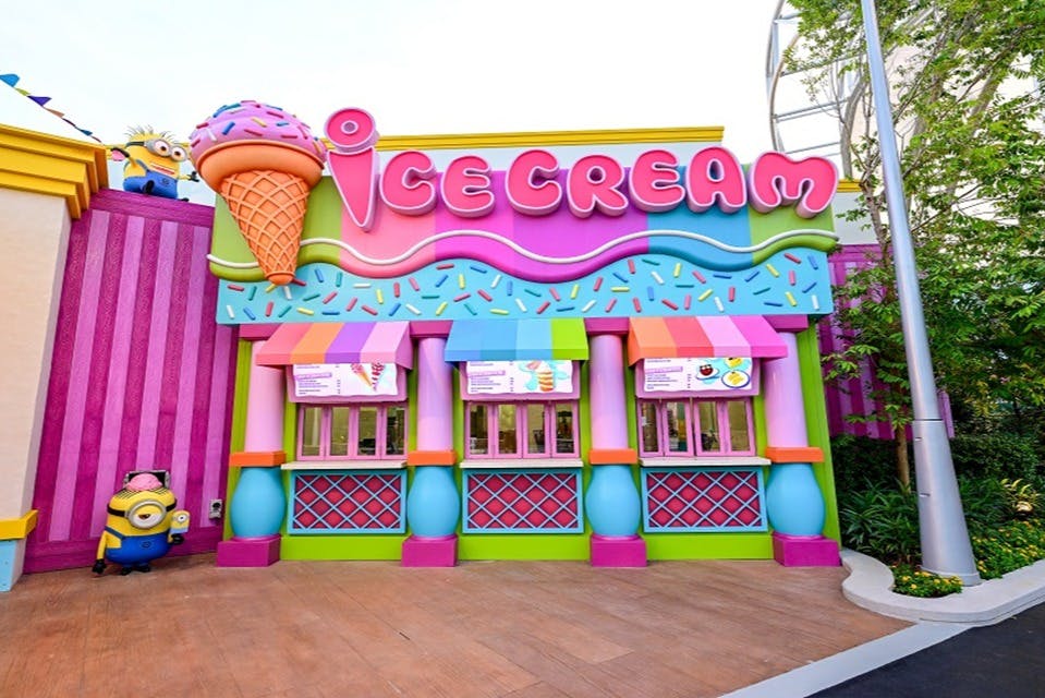 Barevná fasáda zmrzlinářství s výraznými dekoracemi, včetně velkého zmrzlinového kornoutu a zářivých cedulí a markýz.