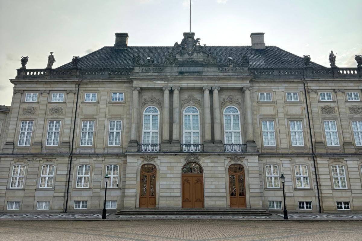 Edificio storico con architettura simmetrica, grandi finestre, dettagli decorati e due porte in legno, visto sotto un cielo nuvoloso.
