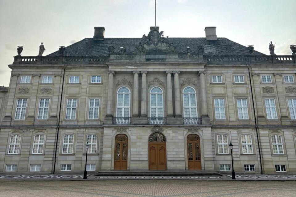 Historic building with symmetrical architecture, large windows, ornate detailing, and two wooden doors, seen under a cloudy sky.