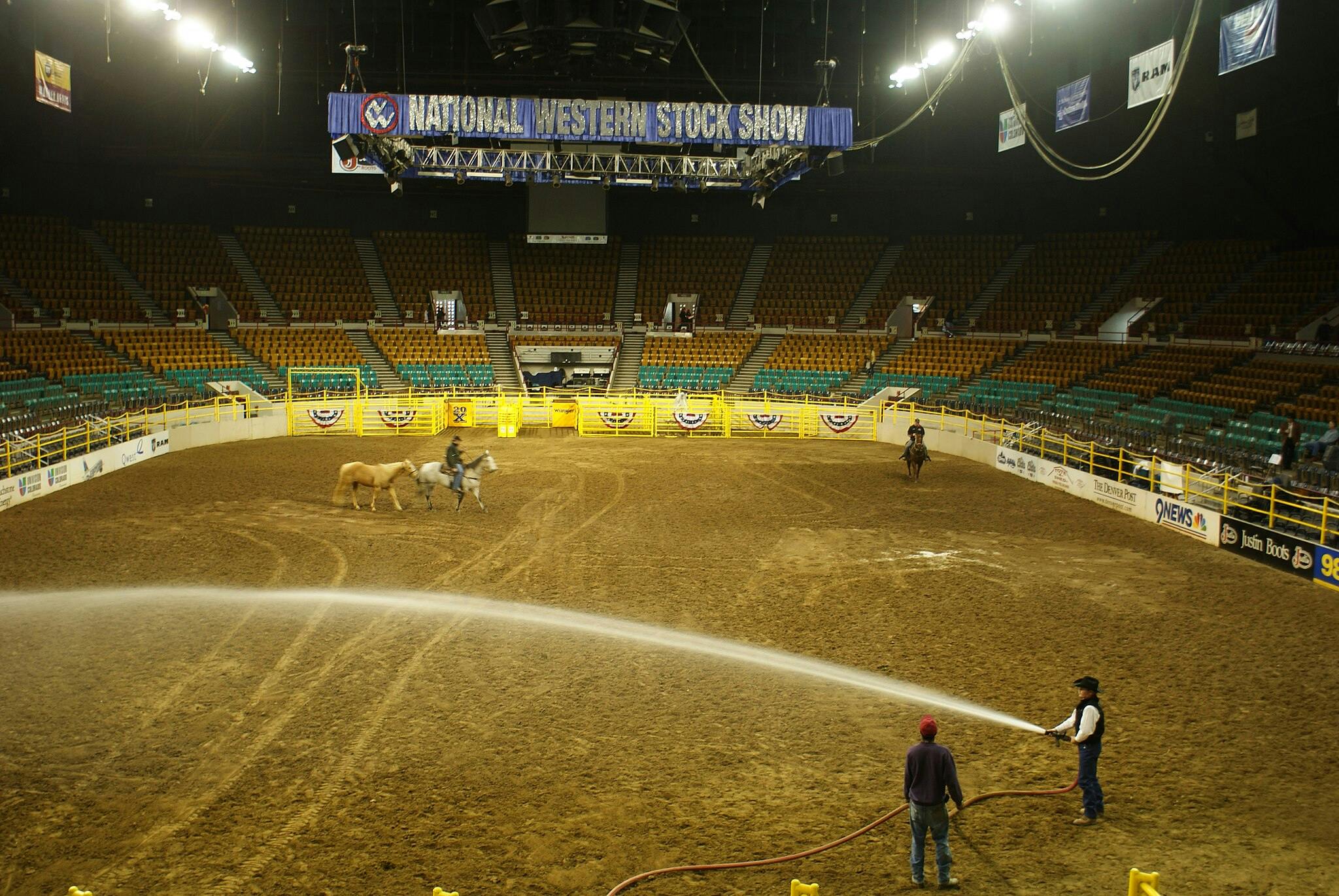 An indoor arena with a "National Western Stock Show" banner, riders on horses, and workers watering the dirt floor.