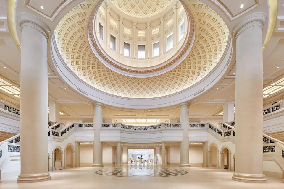 A grand hall with a domed ceiling, intricate designs, and a staircase with ornate railings, illuminated by natural light.