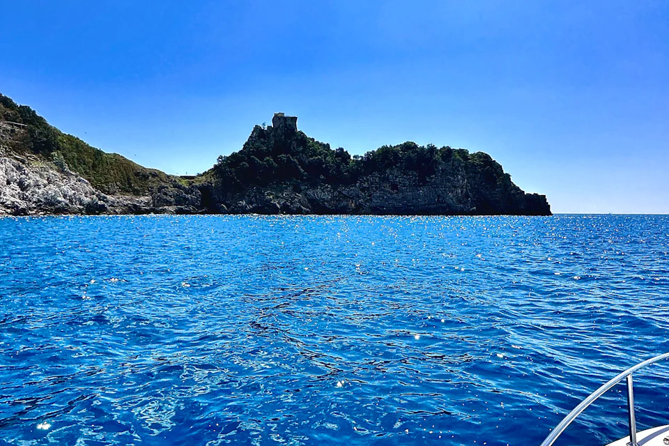 A small island with a rocky shoreline, covered in trees, and a castle on top, viewed from a bright blue sea under a clear sky.