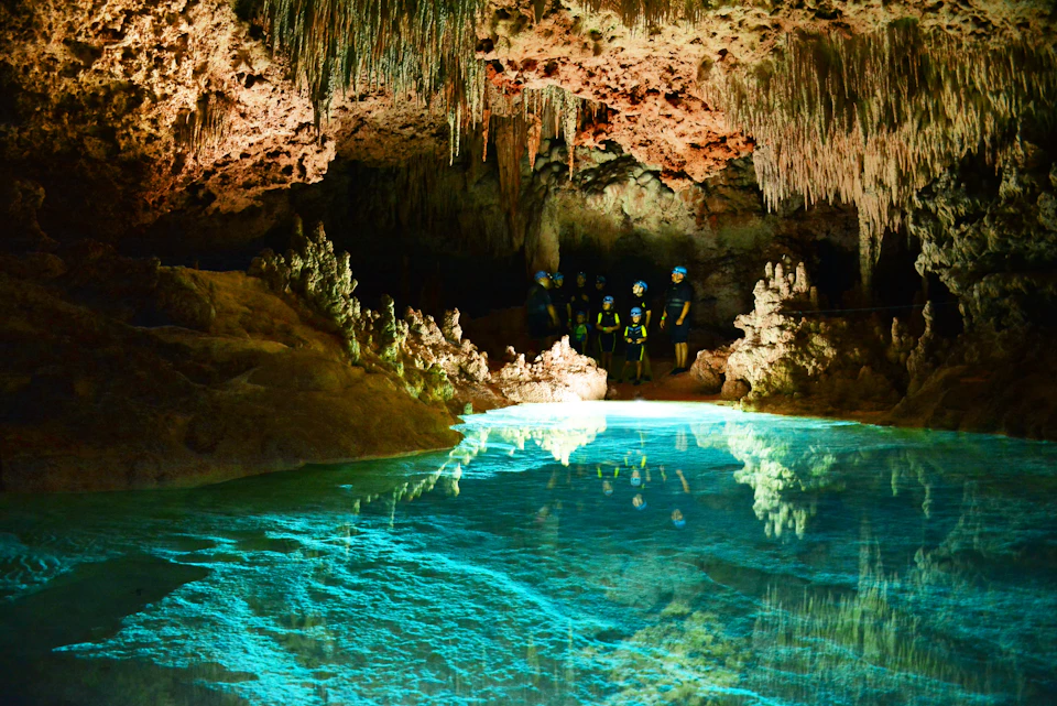 A group of people with helmets stand in an illuminated cave with stalactites, stalagmites, and a pool of clear water.