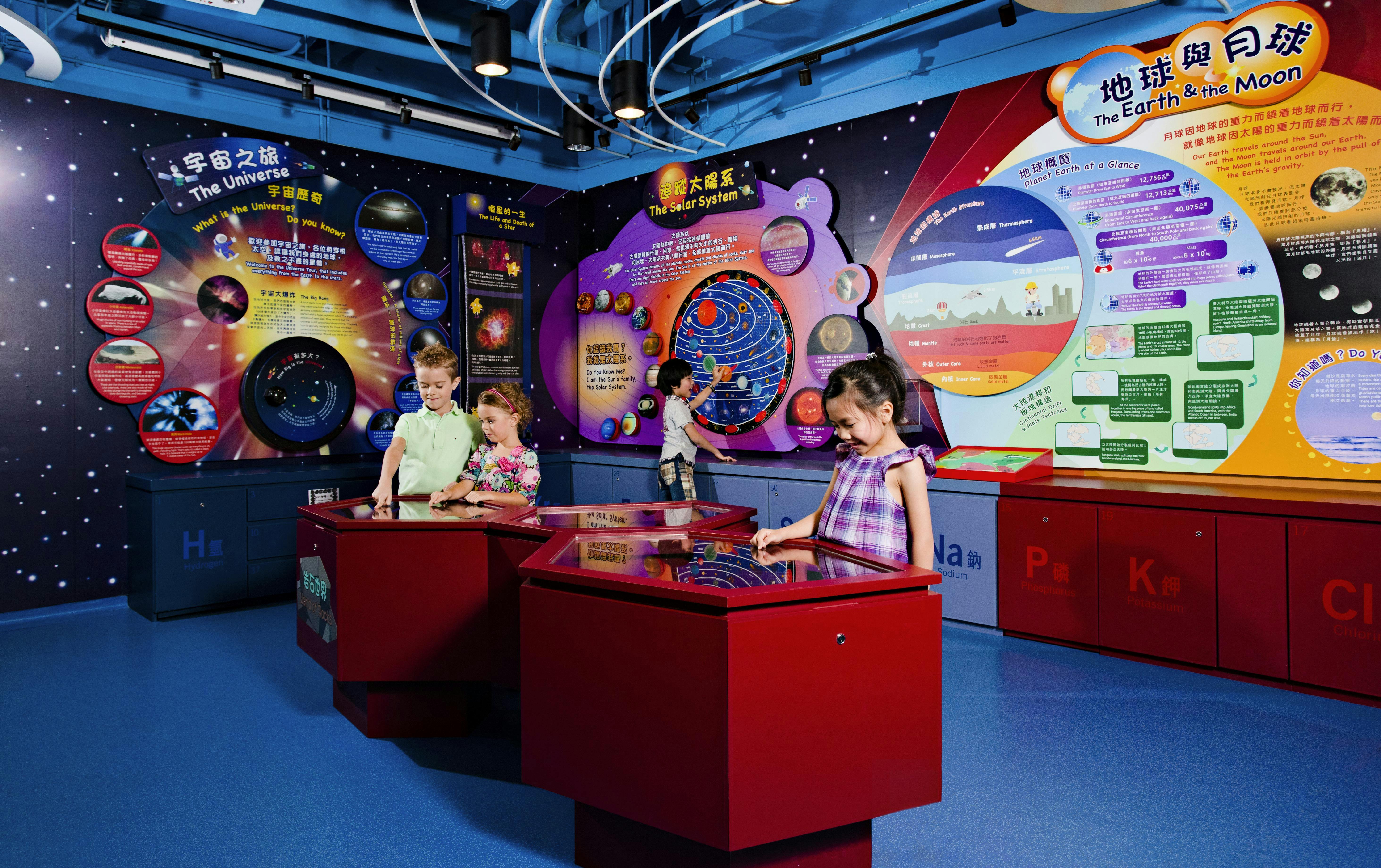 Дети взаимодействуют с красными сенсорными дисплеями на космической тематической выставке с плакатами о Вселенной и Солнечной системе.