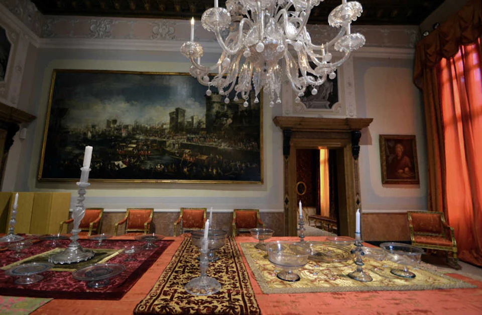 Elegant dining room with a chandelier, ornate tablecloth, silverware, and chairs. A large historic painting hangs on the wall.