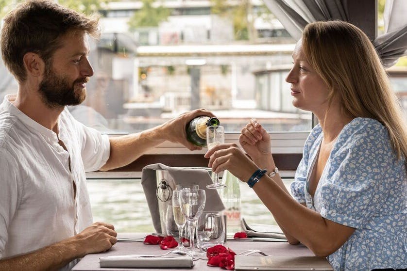Ein Mann im weißen Hemd schenkt Champagner an einem mit Rosenblättern, Gläsern und einer Speisekarte geschmückten Tisch ein, im Hintergrund ist eine Stadtlandschaft zu sehen.