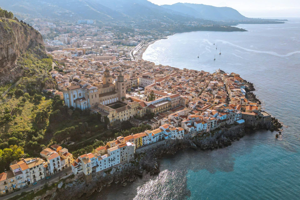 Aerial view of a coastal town with densely packed buildings, a prominent cathedral, and rugged cliffs leading to the sea.