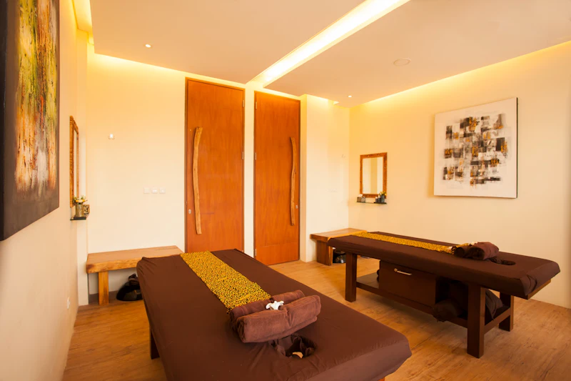 A serene spa room with two massage tables, wooden furniture, warm lighting, and a minimalist decor style.