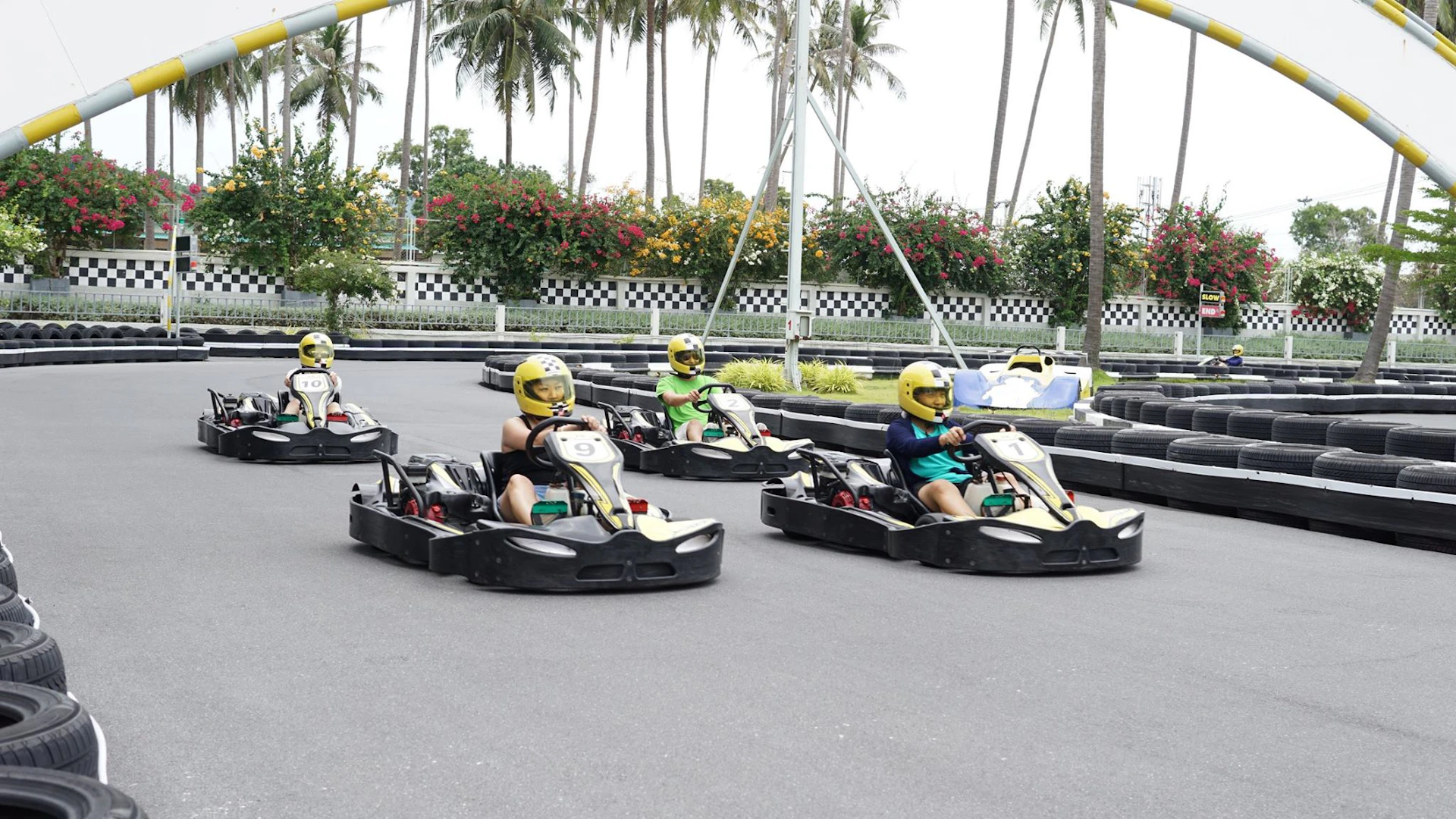 People wearing helmets race go-karts on an outdoor track surrounded by palm trees and greenery.