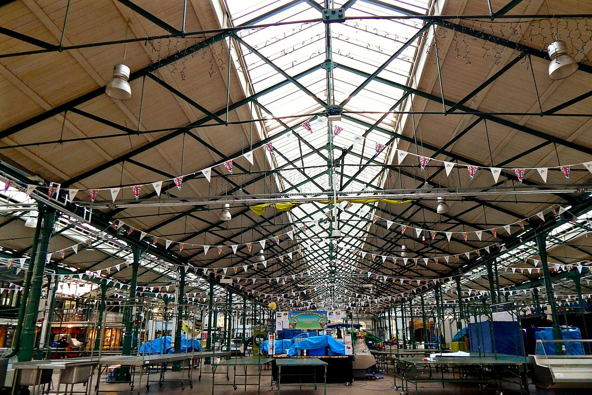 Un mercat interior buit amb un marc metàl·lic verd, banderes, llums penjants i taules cobertes amb lones blaves.