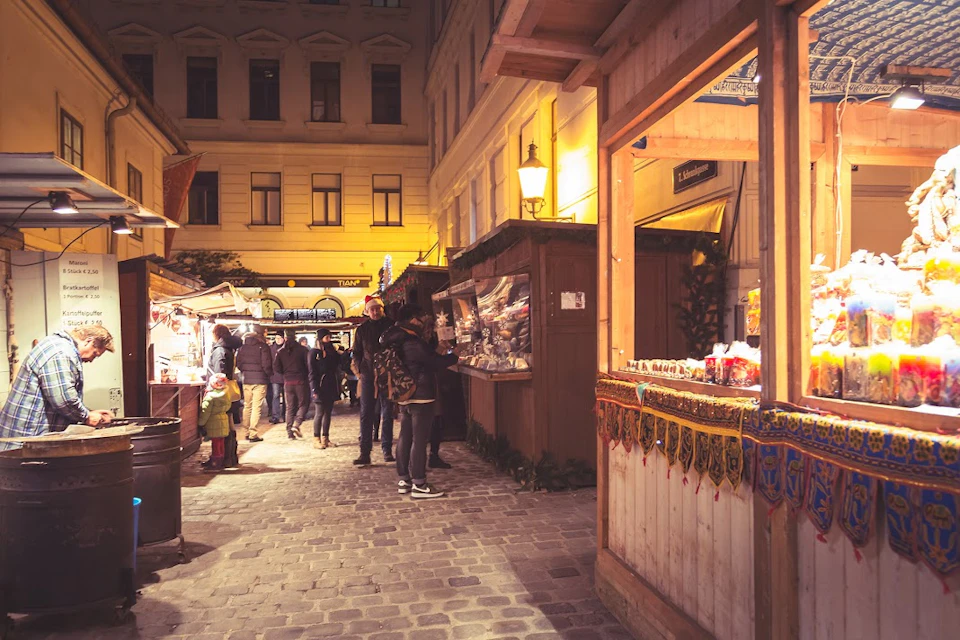People browsing wooden market stalls along a cobblestone street at night, with a warmly lit building in the background.