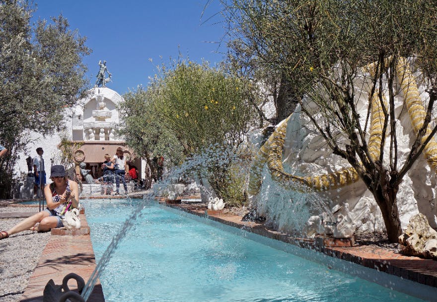 プール、彫像、散水、木々、そして澄んだ青い空の下で背景に座っている人々がいる屋外のシーン。