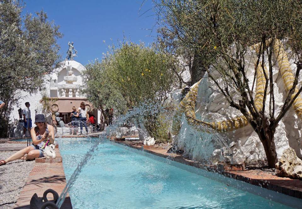 People gather near a turquoise fountain, surrounded by greenery and a decorative snake sculpture, under a clear blue sky.