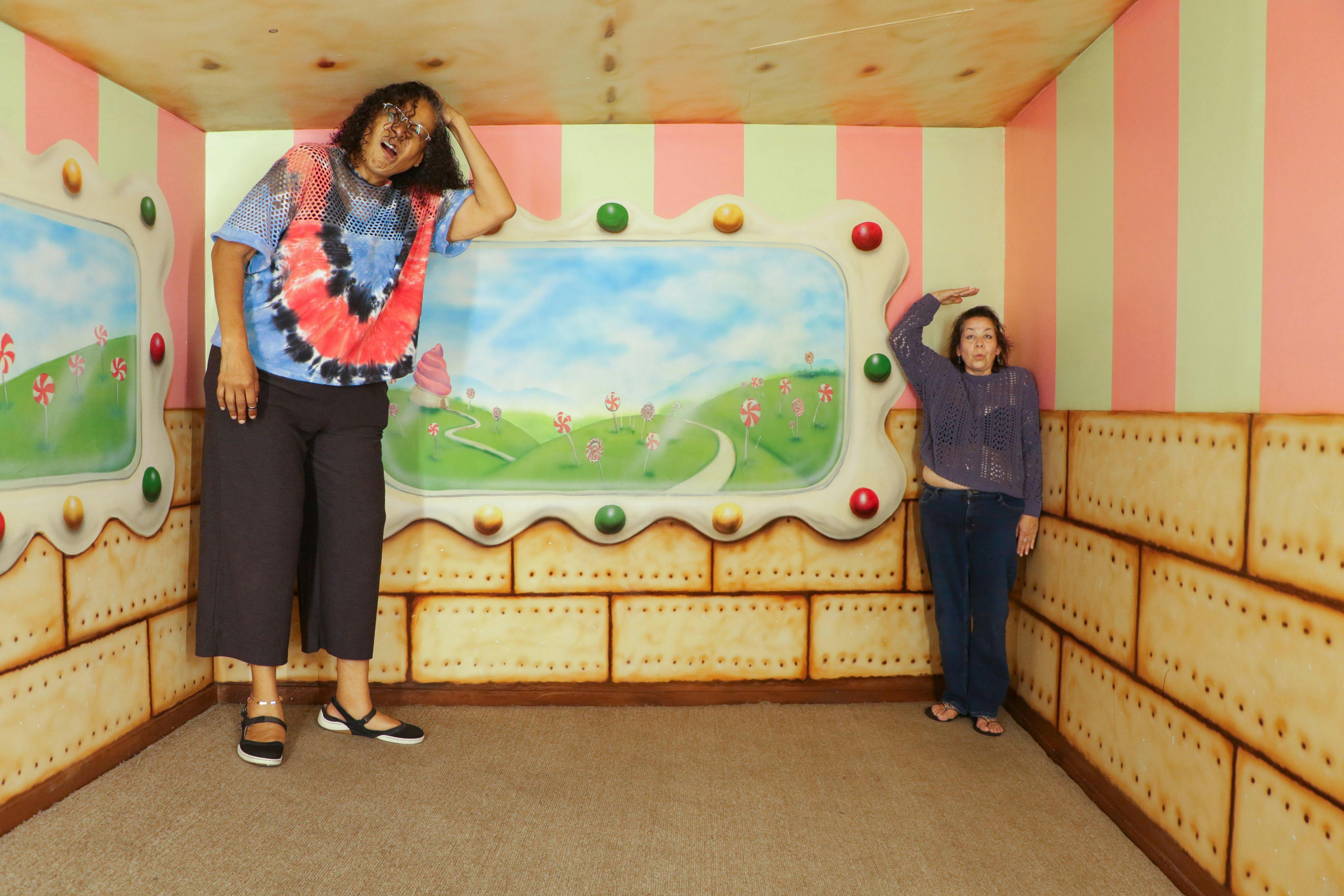 キャンディーをテーマにしたウォールアートが施されたカラフルな部屋にいる2人の女性。1人は天井付近で大きく、もう1人は床付近で小さく見える。