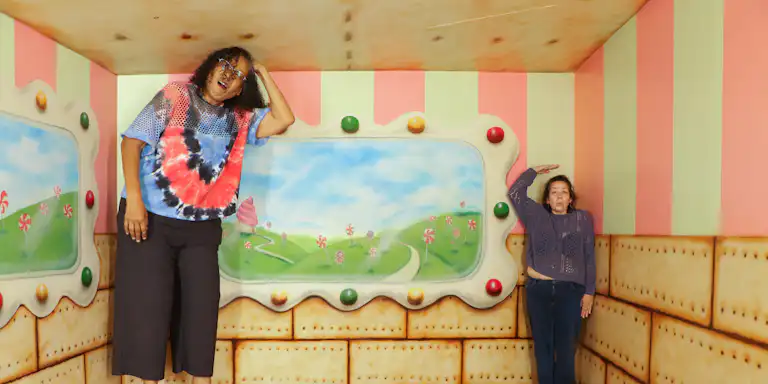 Two women in a colorful room with candy-themed wall art, one appears larger near the ceiling, and the other smaller near the floor.