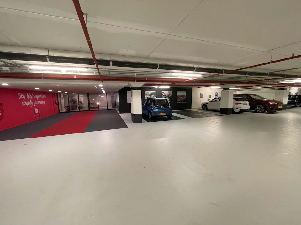 A parking garage with a blue car and two white cars parked. Red and gray carpet leads to a glass double door entry.
